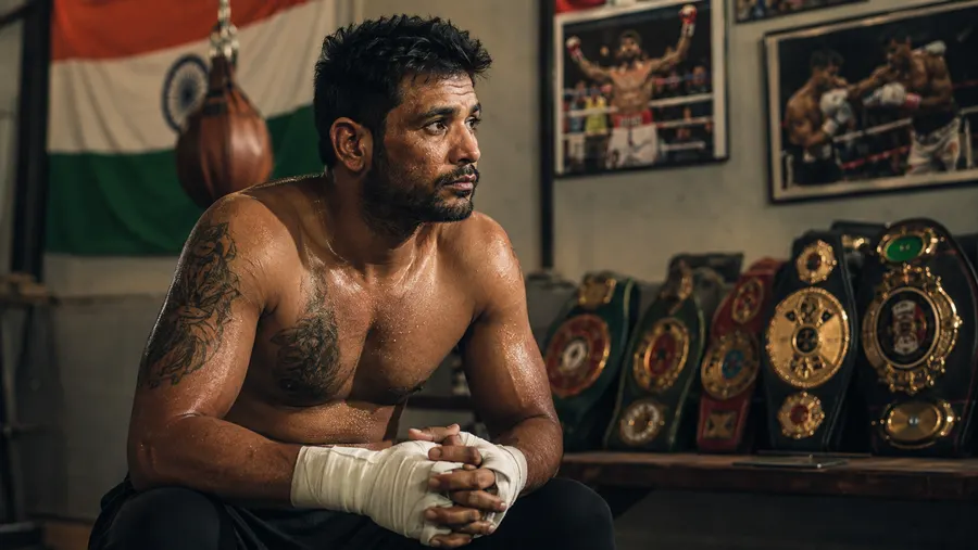 Neeraj Goyat sitting in a gym with his hands wrapped, championship belts displayed behind him
